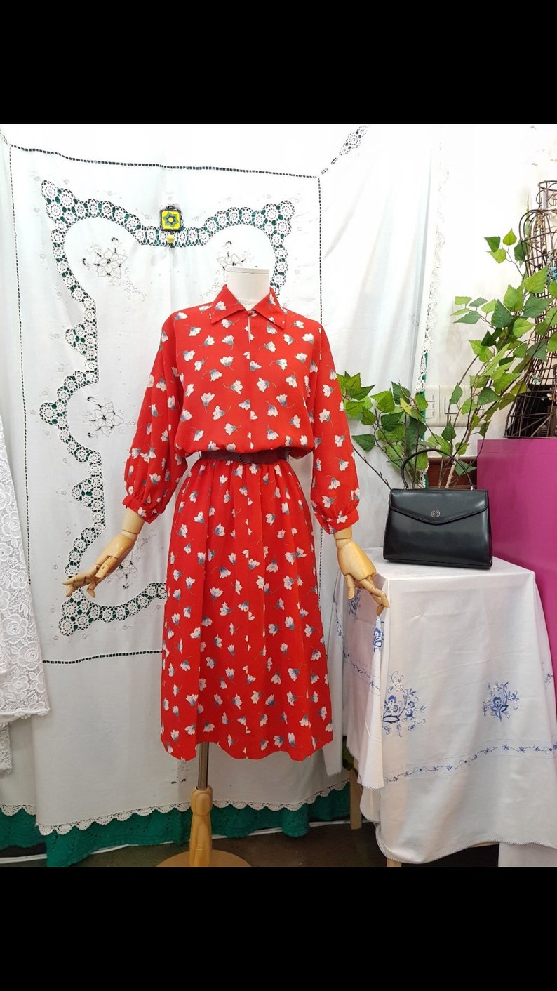 May include: A vintage red dress with a white floral pattern, long sleeves, and a collared neckline. The dress is displayed on a mannequin. A black handbag sits on a table nearby. The dress has a belt at the waist.