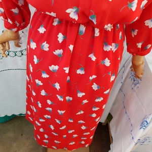 May include: A red dress patterned with white flowers. The dress features long sleeves and a gathered waist. The fabric looks lightweight, and the dress is displayed on a wooden mannequin.