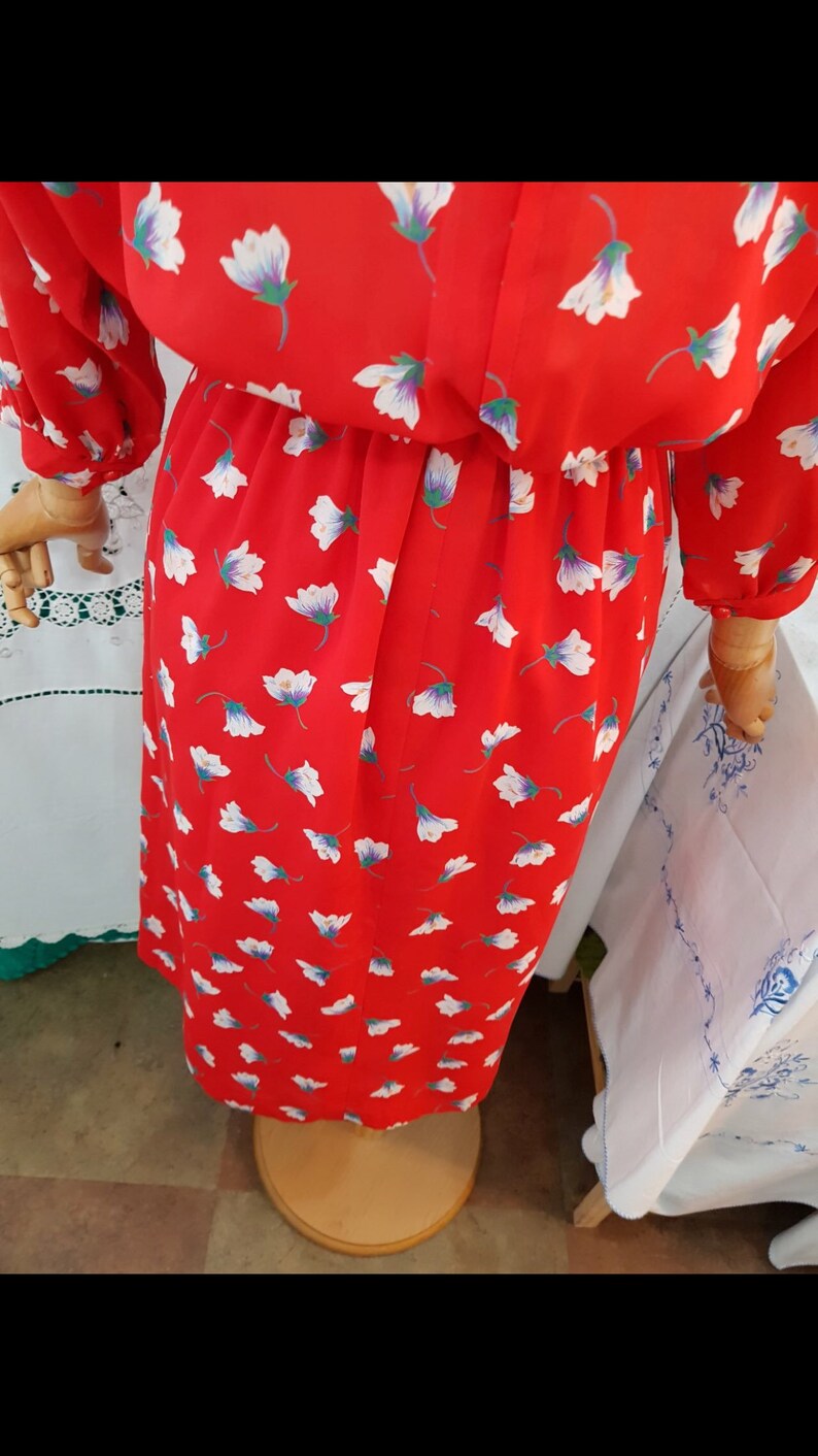 May include: A red dress patterned with white flowers. The dress features long sleeves and a gathered waist. The fabric looks lightweight, and the dress is displayed on a wooden mannequin.