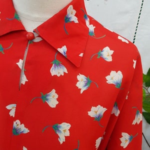 May include: Red shirt with a floral print. The shirt has a repeating pattern of white flowers with purple and green details. A single red button fastens the collar.