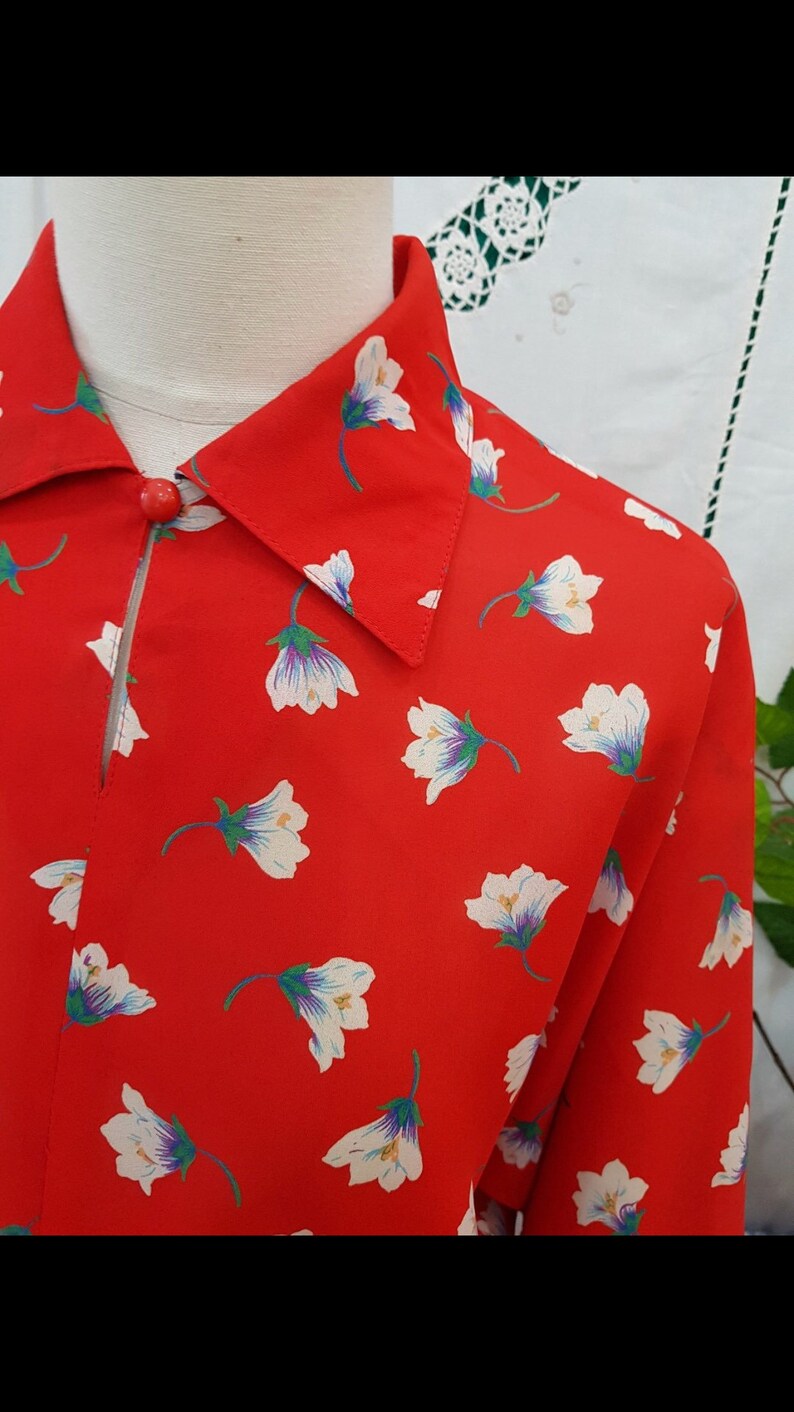 May include: Red shirt with a floral print. The shirt has a repeating pattern of white flowers with purple and green details. A single red button fastens the collar.