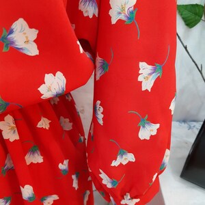 May include: Close-up of a red dress with a floral pattern. The dress features white flowers with purple and green accents. The sleeves are long and gathered at the wrist. The fabric appears to be lightweight.