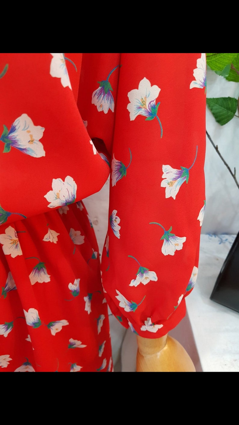May include: Close-up of a red dress with a floral pattern. The dress features white flowers with purple and green accents. The sleeves are long and gathered at the wrist. The fabric appears to be lightweight.