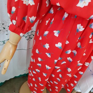 May include: A red dress with a floral pattern of white and purple flowers. The dress has long sleeves and is displayed on a wooden mannequin arm and torso. The dress is on a wooden stand.
