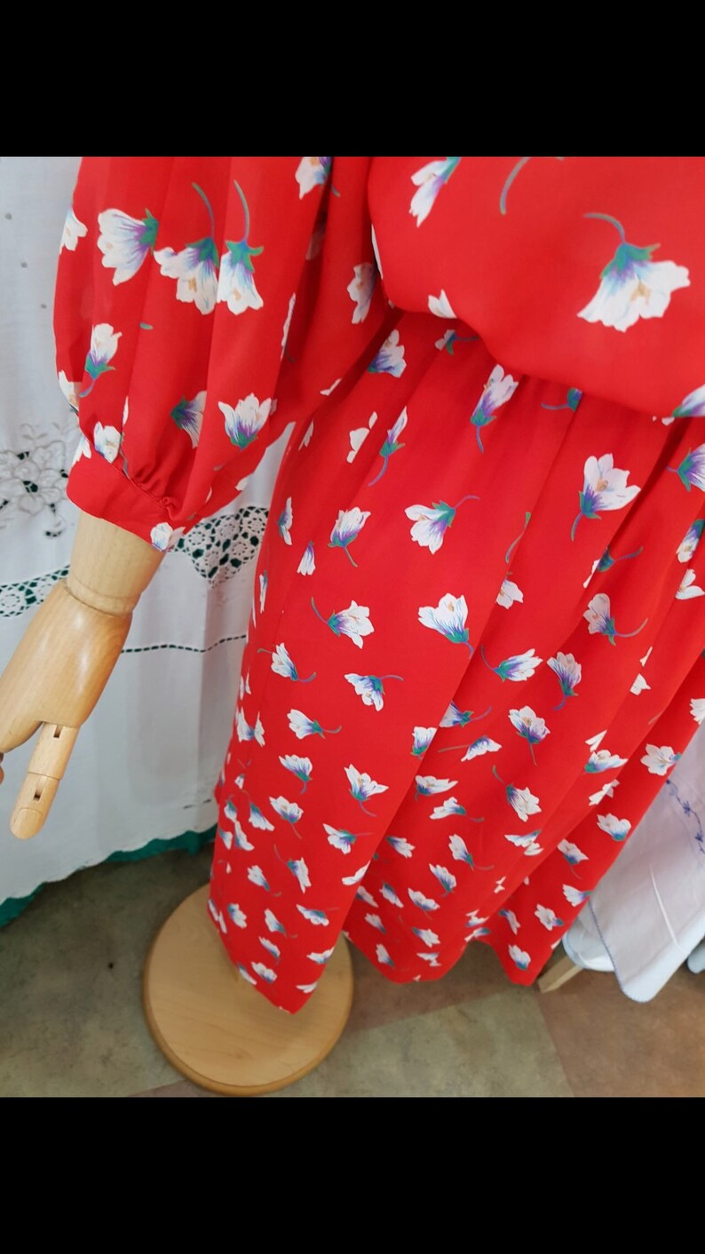 May include: A red dress with a floral pattern of white and purple flowers. The dress has long sleeves and is displayed on a wooden mannequin arm and torso. The dress is on a wooden stand.