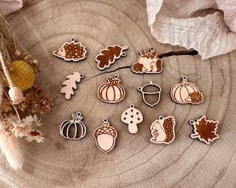 May include: A collection of wooden fall-themed charms, including pumpkins, acorns, leaves, a mushroom, and a squirrel. The charms are laser-cut with intricate details and have small holes for stringing. The wood has a natural, light brown color.