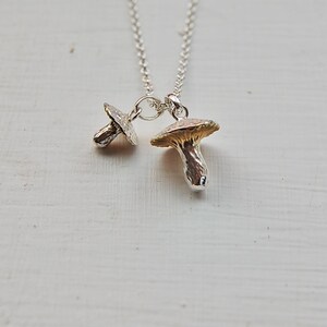 May include: A silver necklace with two mushroom charms. The larger charm is gold-toned and the smaller charm is silver.