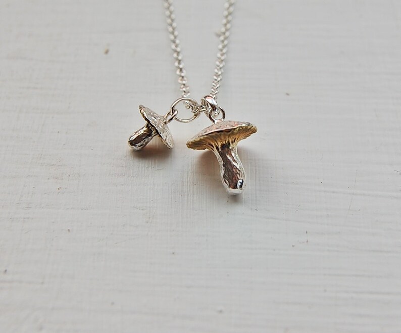 May include: A silver necklace with two mushroom charms. The larger charm is gold-toned and the smaller charm is silver.
