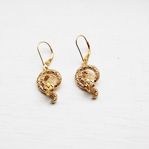 May include: A pair of gold snake earrings with red gemstone eyes. The snakes are coiled and have a textured surface.