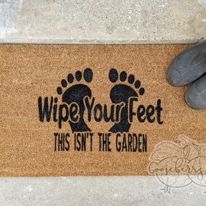 Könnte beinhalten: Eine braune Kokosfaser-Fußmatte mit dem Text "Wipe Your Feet This Isn't The Garden" in schwarzen Buchstaben. Zwei schwarze Fußabdrücke befinden sich über dem Text.