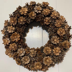 May include: A circular wreath constructed from numerous brown pine cones arranged in a dense, overlapping pattern. The pine cones are oriented to display their flat, flower-like faces, creating a textured, uniform surface across the entire ring.
