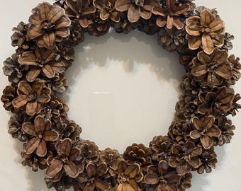 May include: A circular wreath constructed from numerous brown pinecones. The pinecones are arranged in a dense, overlapping pattern, with several individual pinecone scales shaped to resemble flower blossoms distributed around the ring.