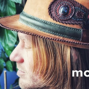 Op de afbeelding: Een bruine leren fedora-hoed met een groene band en een decoratieve veer. De hoed heeft een kralenontwerp en een turquoise steentje. De hoed wordt gedragen door een persoon met lang bruin haar. De tekst "model hat 2" is zichtbaar in de afbeelding.