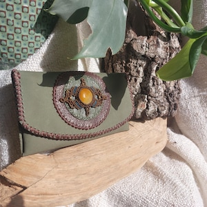 Op de afbeelding: Olijfgroene leren clutch met een decoratief cirkelvormig ornament met een grote oranje edelsteen en ingewikkelde macramé details. De clutch heeft een klepsluiting en bruine stiksels.