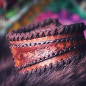 Op de afbeelding: Een bruin leren armband met een decoratief gestikt ontwerp en een donkerbruine suède rand.