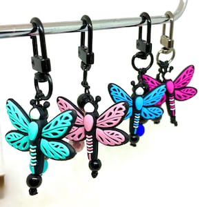 May include: Four dragonfly keychains in a row, each with a different color scheme. The dragonflies have black bodies, white accents, and wings in teal, pink, and blue. The keychains have black clasps and are hanging from a silver bar.