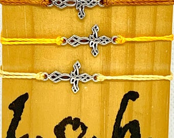 May include: Three adjustable bracelets with silver-colored cross charms on yellow, gold, and orange cords. The bracelets are displayed on a wooden surface with the text "L.O.h." in black script.
