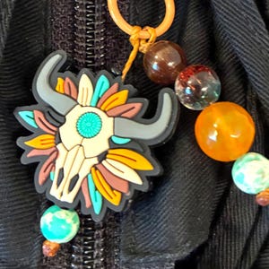 May include: A colorful charm featuring a bull skull design with turquoise, yellow, and brown accents. The charm is attached to a key ring with a collection of decorative beads in shades of brown, orange, and green. The charm is attached to a black fabric.