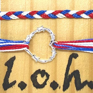 May include: Two bracelets with red, white, and blue braided and striped designs. The bottom bracelet has a silver-colored, heart-shaped charm. The background is a light brown surface with the handwritten letters "L.o.h."