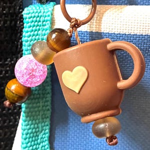 May include: A brown coffee mug keychain with a gold heart charm. The keychain has a series of colorful beads, including pink, brown, and clear. The background is a blue and white striped pattern.