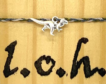 May include: A silver dinosaur charm bracelet with a braided cord in shades of gray, green, and white. The bracelet is displayed on a light brown wooden surface with the letters "L.o.h" written in black.