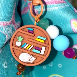 May include: A brown keychain charm shaped like a bookshelf, featuring a sleeping white cat, a small plant, and colorful books. The charm is attached to a light orange clasp and string, with a strand of colorful beads. The background is a teal fabric.