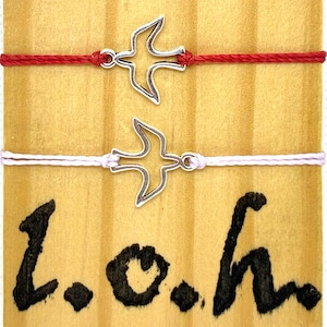 May include: Two friendship bracelets with a silver bird charm. One bracelet is red, the other is white. The bracelets are displayed on a wooden surface with the letters "L.O.h" in black.