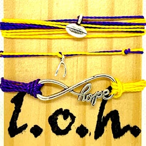 May include: Three handmade bracelets with purple and yellow cords. One bracelet has a football charm, another has a wishbone charm, and the third has an infinity symbol with the word "hope". The background is a wooden surface with the letters "L.O.h."