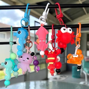 May include: A collection of six colorful sea creature keychains hanging from a black bar. The charms include a light blue seahorse, a teal seahorse, a light purple jellyfish, a pink squid, a red crab, a red lobster, and an orange crab.