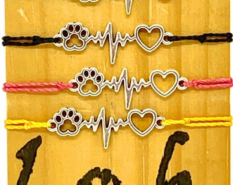 May include: Four colorful string bracelets with silver charms. Each charm features a paw print, a heartbeat, and a heart. The bracelets are arranged in a row on a wooden surface with the text "l.o.h." written below.