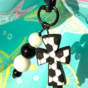 May include: A black and white cross-shaped charm with a soccer ball pattern, attached to a black clasp and a string of black and white beads. The charm hangs against a turquoise background with a floral pattern.