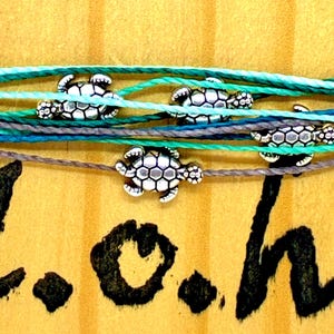 May include: A set of three bracelets made from thin, multi-colored cords in shades of teal, turquoise, and grey. Each bracelet features a small, silver-toned metal sea turtle charm. The text L.o.h. is written in black ink below the bracelets.