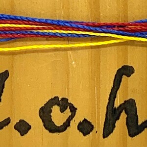 May include: A close-up of a wooden surface with a yellow, red, and blue string tied around it. The text "L.o.h." is written in black paint on the surface.