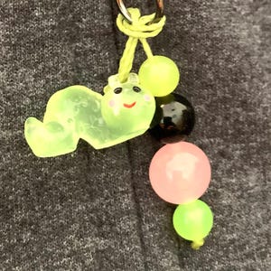 May include: A whimsical keychain featuring a translucent green caterpillar charm with a smiling face and pink cheeks. The charm is attached to a string with a black, pink, and green bead design, and a silver-colored clasp.