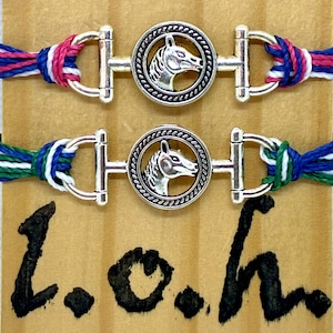 May include: Two friendship bracelets with silver-colored horse head charms. One bracelet has a red, white, and blue braided cord, and the other has a green, white, and blue braided cord. The bracelets are on a light brown surface with the letters "L.o.h" in black.