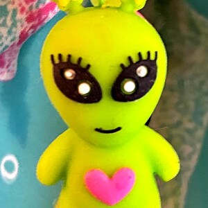 May include: A lime green alien charm with black eyes, eyelashes, and a pink heart on its chest. The charm has yellow star-shaped accents and a black bead at the bottom, attached to a pink loop.