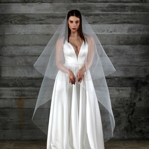 May include: A white satin wedding dress with a plunging neckline and a flowing skirt. A sheer, white veil drapes over the shoulders. The dress is floor-length and has a simple, elegant design, perfect for a wedding.