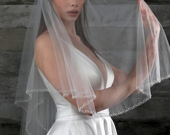 May include: A single-tier bridal veil with a delicate beaded trim. The veil is made of soft tulle and is worn by a woman with dark hair.