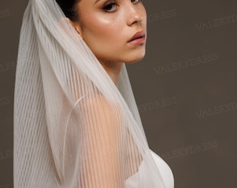 May include: A sheer white veil with vertical pleats, draped over the head and shoulders. The veil is paired with a white dress. The image is set against a neutral background, creating a soft, elegant look. The veil's texture adds visual interest.