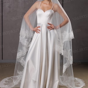 May include: A white satin wedding dress with a sweetheart neckline and spaghetti straps. The dress has a flowing A-line skirt and is paired with a long, sheer veil trimmed with lace. The veil drapes over the shoulders.