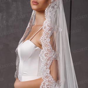May include: A white bridal veil with intricate lace detailing along the edges. The veil is draped over a white satin wedding dress. The image is set against a neutral background, highlighting the delicate design of the veil.