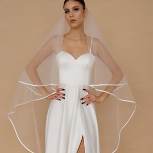 May include: A white wedding dress with a sweetheart neckline and spaghetti straps. The dress has a high slit and is paired with a sheer veil with a white trim. The background is a solid tan color.