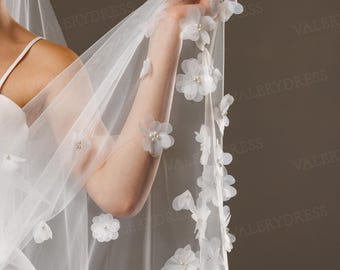 May include: A white bridal veil adorned with delicate, three-dimensional fabric flowers and small pearl accents. The veil is sheer and flowing, with the floral embellishments clustered along the edge. The overall effect is elegant and romantic.