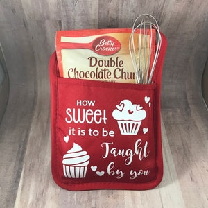 May include: Red oven mitt with a pocket for a whisk and a package of chocolate chip cookie mix. The oven mitt has a white design that says "How sweet it is to be taught by you" with two cupcake illustrations.