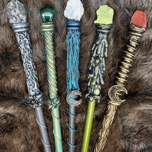K&ouml;nnte beinhalten: F&uuml;nf handgefertigte Zauberst&auml;be mit einzigartigen Designs und Edelsteinakzenten. Jeder Zauberstab hat ein anderes Farbschema, darunter Silber, Gr&uuml;n, Blau und Gold, mit aufwendigen Details und dekorativen Elementen wie einer Spinne und einem Mond.