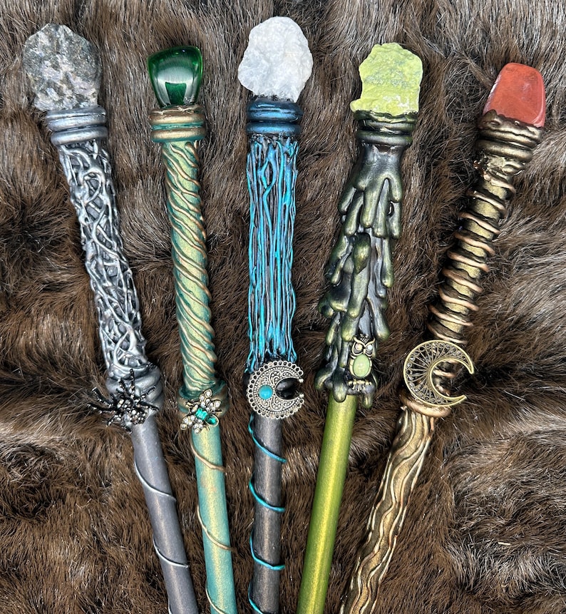 K&ouml;nnte beinhalten: F&uuml;nf handgefertigte Zauberst&auml;be mit einzigartigen Designs und Edelsteinakzenten. Jeder Zauberstab hat ein anderes Farbschema, darunter Silber, Gr&uuml;n, Blau und Gold, mit aufwendigen Details und dekorativen Elementen wie einer Spinne und einem Mond.
