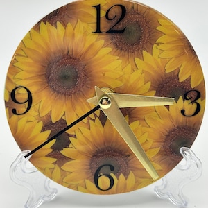 May include: A round clock with a sunflower design. The clock face has black numbers and gold hands. The clock is on a clear plastic stand.