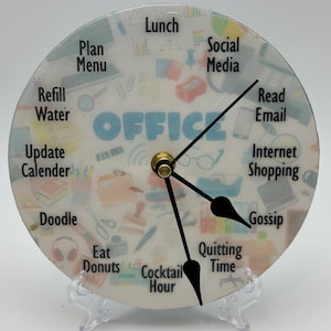 May include: A white clock with black hands and a clear face. The face is decorated with office-themed illustrations and the word "OFFICE" in blue. The clock is on a clear plastic stand.