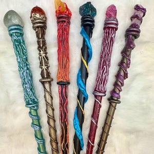 K&ouml;nnte beinhalten: Eine Sammlung von sieben handgefertigten Zauberst&auml;ben mit einzigartigen Designs. Jeder Zauberstab hat ein anderes Farbschema, darunter Gr&uuml;n, Braun, Rot, Schwarz und Lila, und ist mit einem Kristall oder Edelstein gekr&ouml;nt. Die Zauberst&auml;be weisen komplizierte Details und Spiralmuster auf.