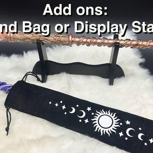 K&ouml;nnte beinhalten: Ein kupferfarbener Zauberstab mit Spiralmuster, ausgestellt auf einem schwarzen St&auml;nder. Eine schwarze Zauberstabtasche mit einem Himmelsdesign mit Monden, Sternen und einer Sonne ist ebenfalls vorhanden. Der Text "Add ons: Wand Bag or Display Stand" steht oben.